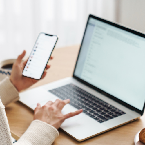 Tischplatte mit einem Laptop, der gerade mit einer Hand einer nicht sichtbaren Person bedient wird. In der anderen Hand hält die Person ein Smartphone.