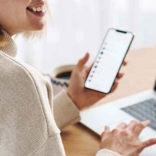 Foto einer Person, die ein Smartphone in der linken Hand hält. Im Hintergrund ist ein Teil eines Laptops zu sehen.